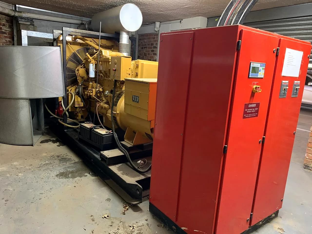 Large standby generator with a yellow diesel engine and alternator in a plant room, beside red switchgear panels
