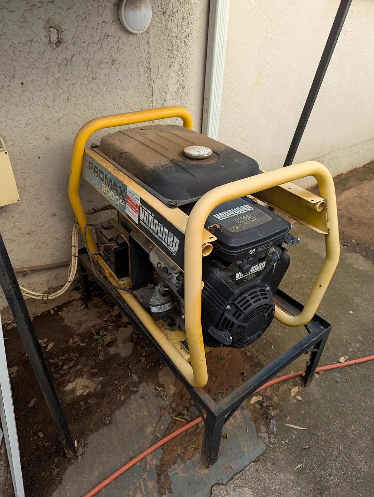 Open-frame petrol generator with a Vanguard engine, on a steel stand against a house wall
