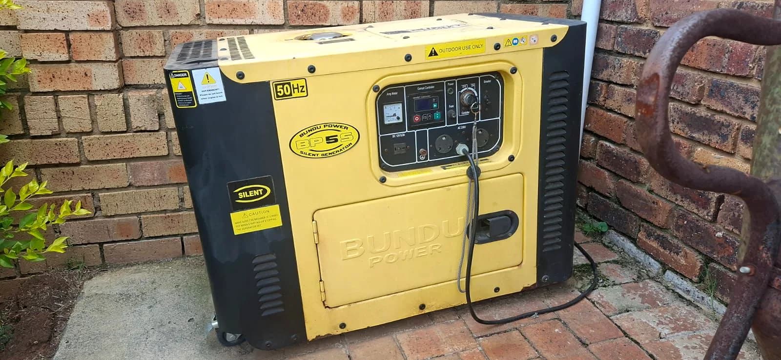 Yellow Bundu Power silent diesel generator with its control panel, against a face-brick wall