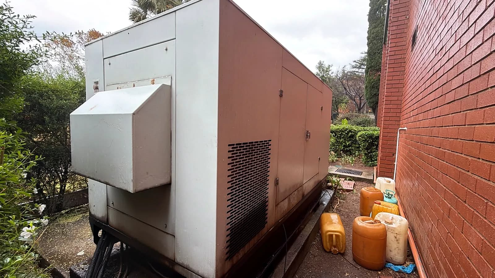 Large generator canopy with rust and dark stains along its base, beside plastic fuel containers stored against a brick wall