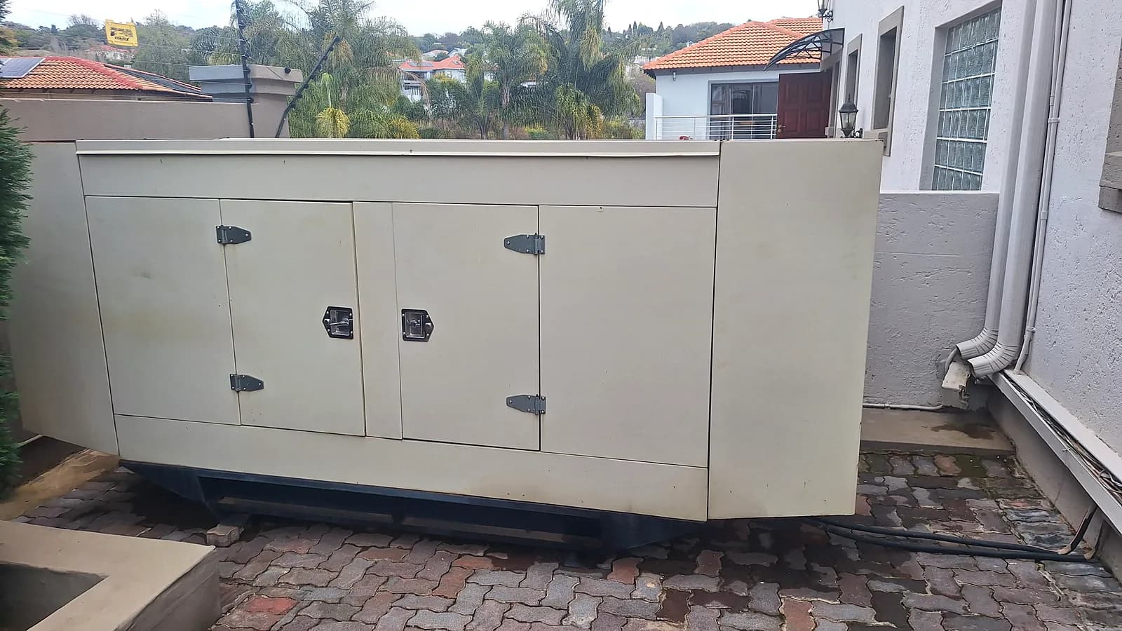 Large beige standby generator on the paved side yard of a house, with neighbouring homes and palm trees behind