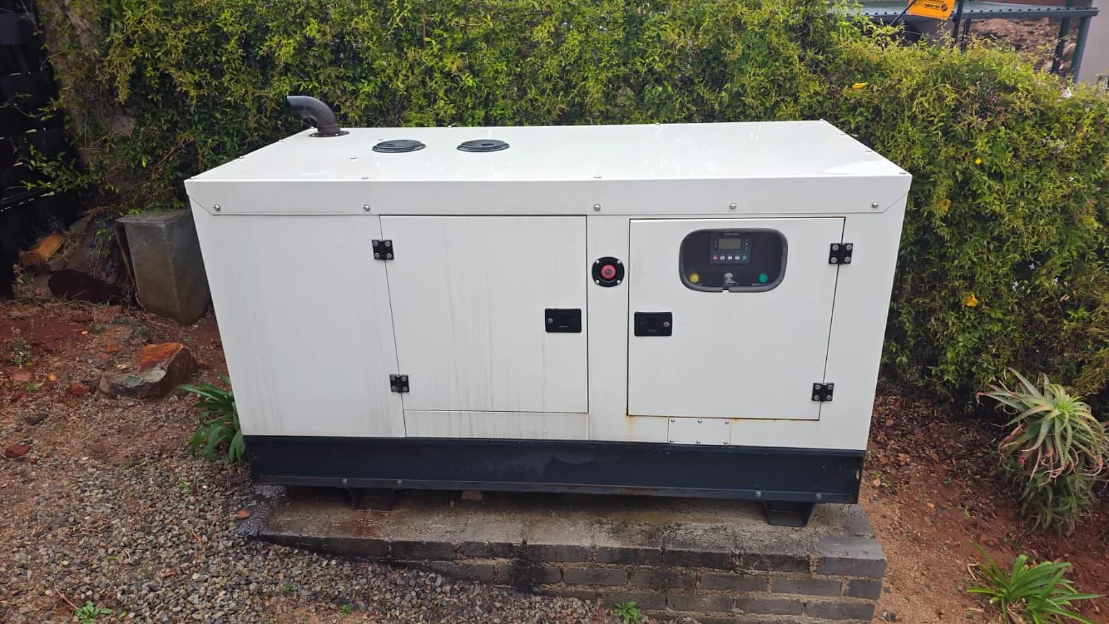 White silent diesel generator with its controller window, standing on a brick plinth in a garden bed