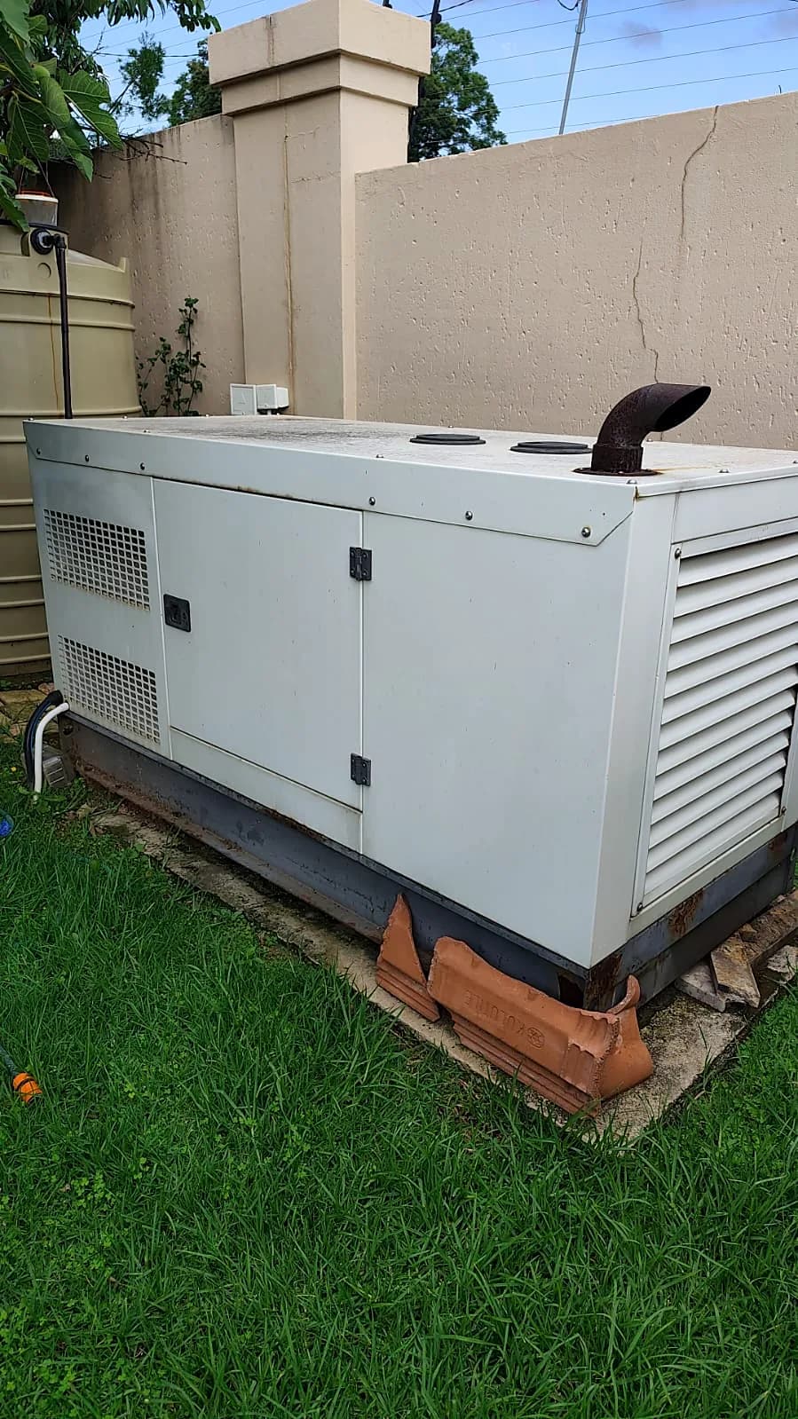 Silent diesel generator on a lawn beside a boundary wall, with a rusty exhaust outlet on its roof