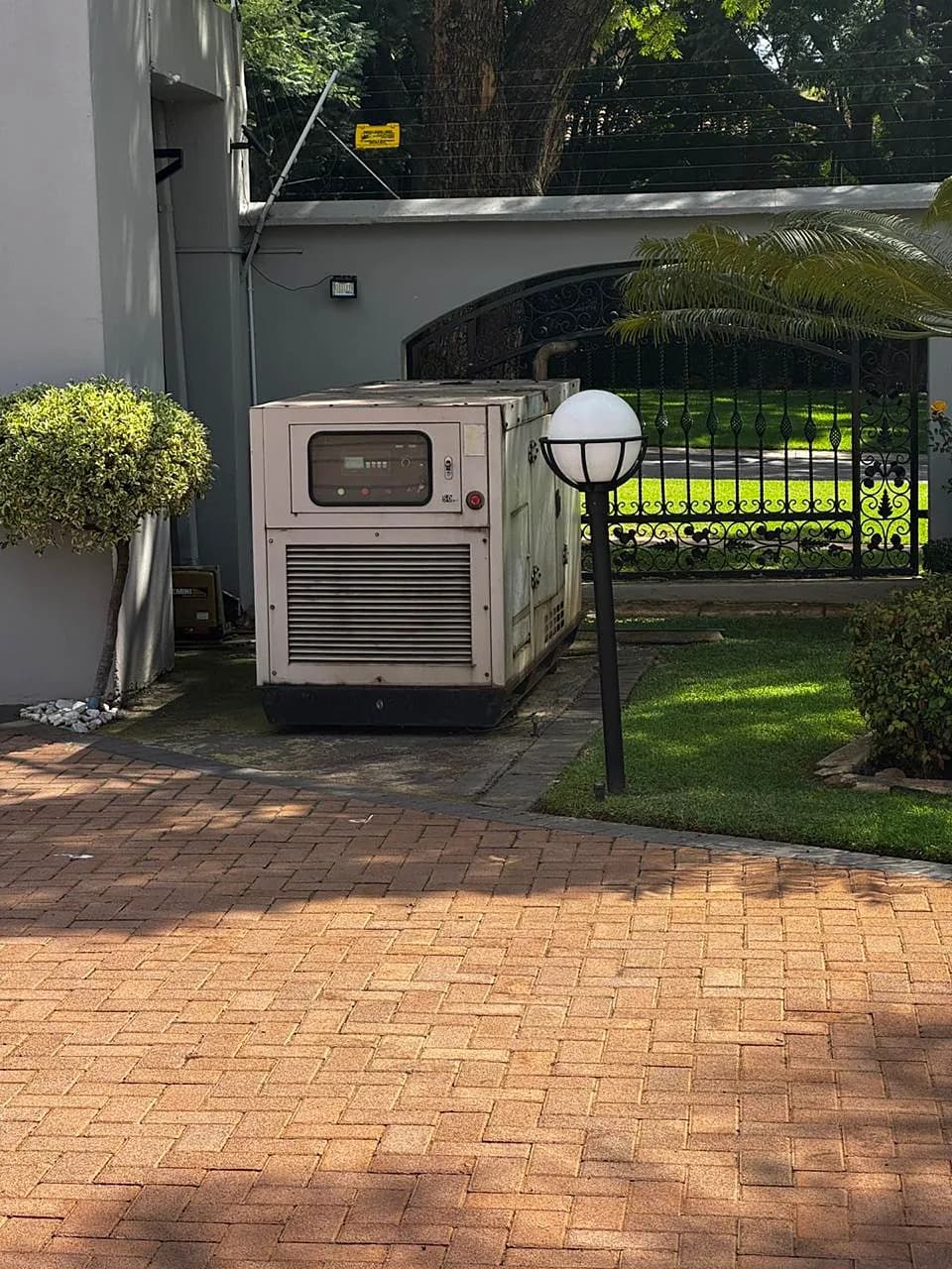 Canopied standby generator beside the paved driveway of a large home, next to a garden lamp and an ornate gate
