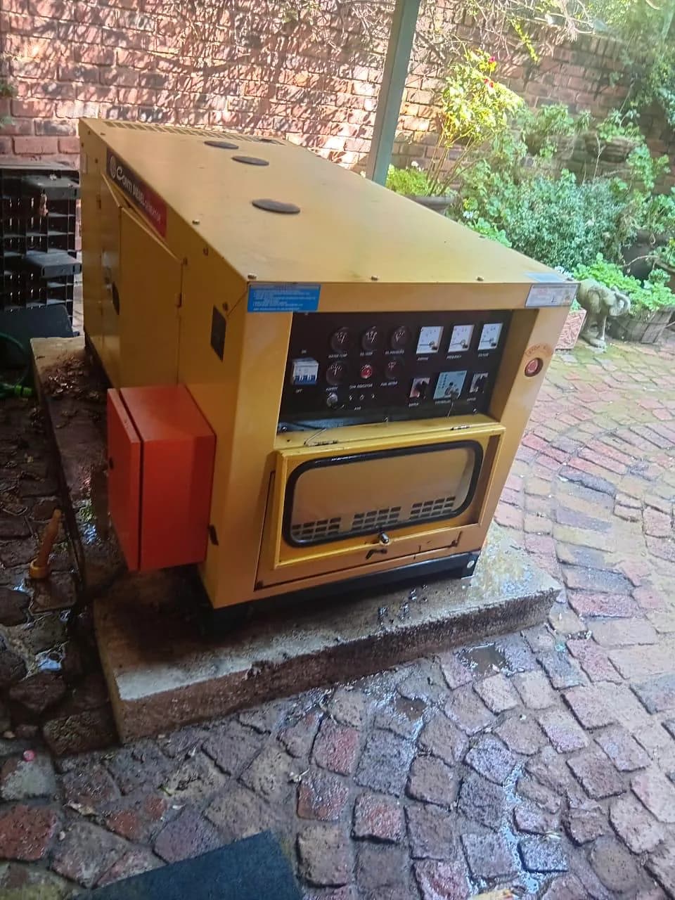 Yellow canopied diesel generator with its instrument panel, on a concrete base beside wet cobbled paving