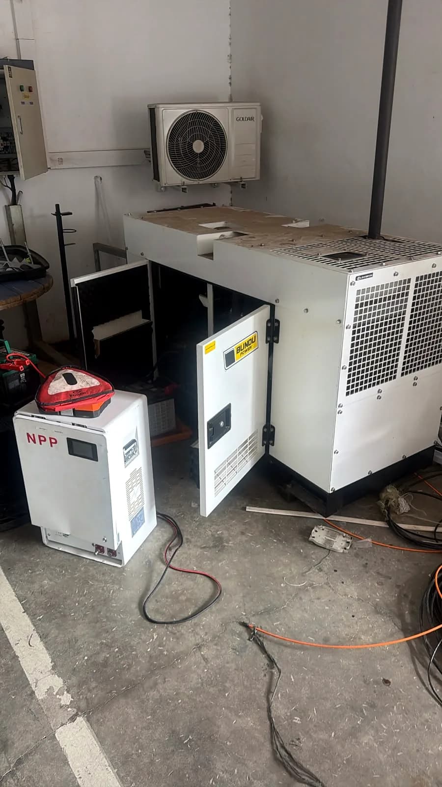 Silent diesel generator with its service doors open in a garage, beside a battery cabinet, with cables on the floor