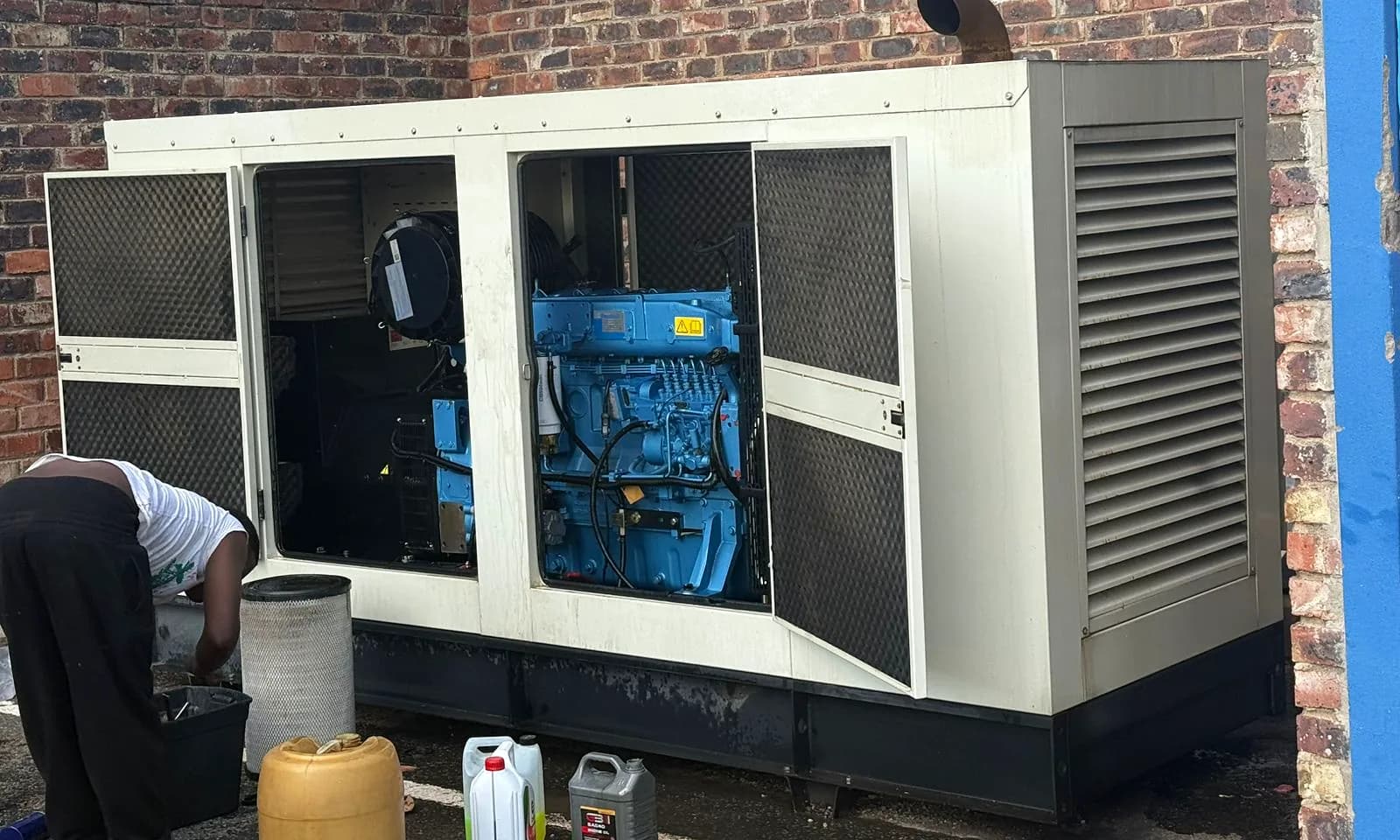 Technician servicing a large diesel standby generator, with the canopy doors open, the blue engine exposed and oil containers on the ground